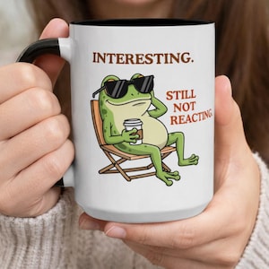 May include: White ceramic mug with a black interior and handle. The mug features a cartoon frog wearing sunglasses and holding a coffee cup, lounging in a chair. Text on the mug reads "INTERESTING." and "STILL NOT REACTING."