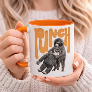 May include: White ceramic mug with an orange interior and handle. The mug features the word "PUNCH" in orange block letters and a graphic of two monkeys hugging. The mug is held by a person.