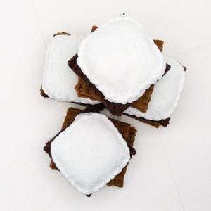 K&ouml;nnte beinhalten: Ein Stapel Filz-Marshmallow-S'mores, jede bestehend aus zwei braunen Quadraten und einem wei&szlig;en Marshmallow in der Mitte. Die S'mores sind in einem Stapel angeordnet, wobei die wei&szlig;en Marshmallows nach oben zeigen.