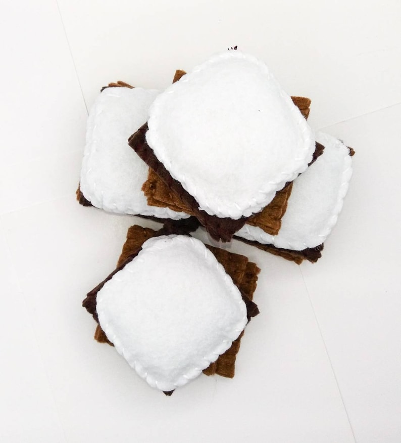 K&ouml;nnte beinhalten: Ein Stapel Filz-Marshmallow-S'mores, jede bestehend aus zwei braunen Quadraten und einem wei&szlig;en Marshmallow in der Mitte. Die S'mores sind in einem Stapel angeordnet, wobei die wei&szlig;en Marshmallows nach oben zeigen.