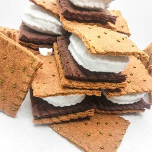 K&ouml;nnte beinhalten: Ein Stapel aus Filz-Graham-Crackern, Marshmallow-Fluff und Schokoladenquadraten, die einem S'more &auml;hneln. Die Filzteile sind zusammengen&auml;ht, um ein realistisches Aussehen zu erzeugen.