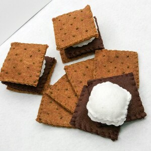 K&ouml;nnte beinhalten: Ein Set aus Filz-S'mores, jede bestehend aus zwei Graham-Cracker-Quadraten und einer wei&szlig;en Marshmallow-F&uuml;llung. Die S'mores sind in einem Stapel angeordnet, wobei sich einige der Graham-Cracker &uuml;berlappen.