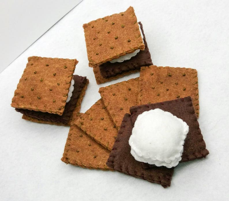 K&ouml;nnte beinhalten: Ein Set aus Filz-S'mores, jede bestehend aus zwei Graham-Cracker-Quadraten und einer wei&szlig;en Marshmallow-F&uuml;llung. Die S'mores sind in einem Stapel angeordnet, wobei sich einige der Graham-Cracker &uuml;berlappen.