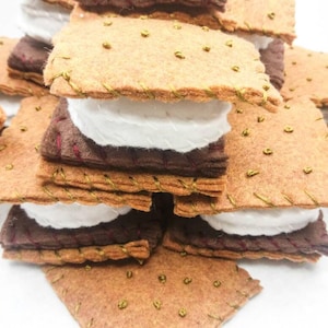 K&ouml;nnte beinhalten: Ein Stapel aus Filz-Graham-Crackern, Schokolade und Marshmallow-Quadraten, die einem S'more &auml;hneln, mit goldenen N&auml;hten an den R&auml;ndern.