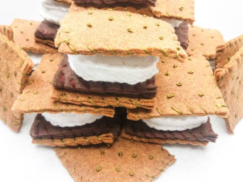 K&ouml;nnte beinhalten: Ein Stapel aus Filz-Graham-Crackern, Schokolade und Marshmallow-Quadraten, die einem S'more &auml;hneln, mit goldenen N&auml;hten an den R&auml;ndern.