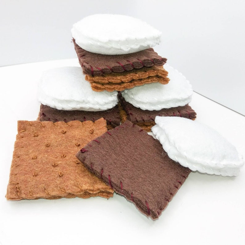 K&ouml;nnte beinhalten: Ein Stapel Filz-S'mores, bestehend aus Graham-Crackern, Schokoladenquadraten und Marshmallows. Die Filzteile sind mit rotem Faden zusammengen&auml;ht.