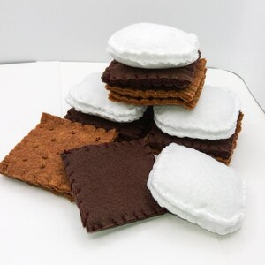 K&ouml;nnte beinhalten: Ein Stapel Filz-S'mores, bestehend aus Graham-Crackern, Schokoladenquadraten und Marshmallows. Die Filzteile sind mit rotem Faden zusammengen&auml;ht.