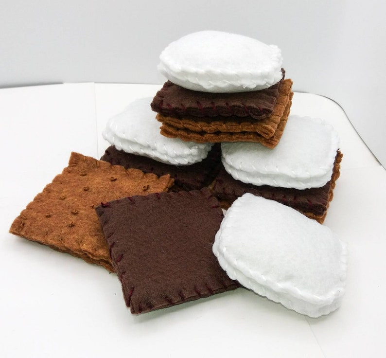 K&ouml;nnte beinhalten: Ein Stapel Filz-S'mores, bestehend aus Graham-Crackern, Schokoladenquadraten und Marshmallows. Die Filzteile sind mit rotem Faden zusammengen&auml;ht.