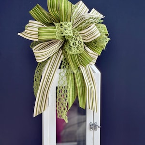 May include: A decorative bow featuring green and white striped ribbon, accented with a delicate green lace. The bow is atop a white, vintage-style lantern with a glass door and metal latch, set against a dark blue background.