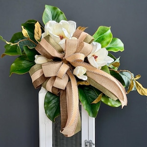 May include: A white floral arrangement with large, creamy white magnolia blossoms and vibrant green leaves. The arrangement is adorned with a multi-toned brown and green ribbon bow. The backdrop is a dark grey surface.