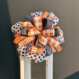 May include: A decorative bow made of wired ribbon featuring a mix of patterns including black and white polka dots, orange and white buffalo plaid, and black and white grid prints. The bow is constructed with multiple loops and long tails.