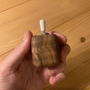 May include: A small, square wooden toggle switch with a white tip. The switch is held in a hand, showcasing the natural wood grain. The toggle switch is a component often used in electronics and DIY projects.