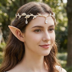 May include: A woman wearing a gold-colored leaf-design headband with dangling teardrop-shaped crystals. She also has prosthetic elf ears. The headband is adorned with small clear crystals. The background is blurred, with green foliage.