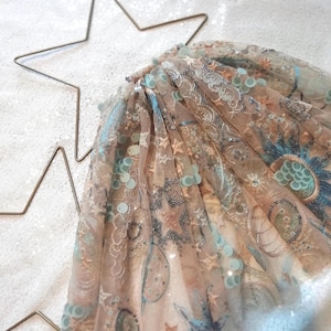 May include: A sheer, pleated skirt made of beige tulle fabric featuring intricate embroidery of blue and silver celestial motifs, including stars and suns, accented with light blue sequins throughout the layered, semi-transparent material.