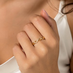 May include: A gold ring featuring an infinity-style twisted band design. The surface of the metal band is embellished with a series of small, clear, sparkling stones set along the interlocking loops, creating a continuous, delicate, and refined appearance.