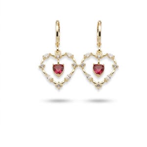 May include: A pair of gold-colored hoop earrings. Each earring features a heart-shaped design with a red heart-shaped gemstone in the center. The heart is surrounded by small, clear gemstones. The earrings are on a white background.