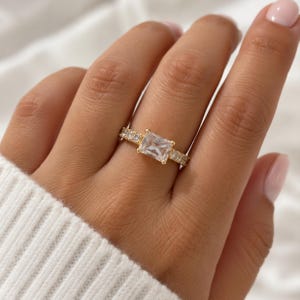 May include: A gold ring featuring a large rectangular emerald-cut clear stone at the center, flanked by a band encrusted with smaller round clear stones. The metal band has a polished gold finish and a prong setting for the main stone.