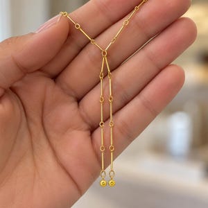 Könnte beinhalten: Eine goldfarbene Halskette mit einer feinen Kette aus kleinen runden Gliedern und länglichen Stabsegmenten. Das Design teilt sich in zwei parallele vertikale Stränge, die jeweils in einem kleinen, runden, texturierten goldfarbenen Anhänger enden.