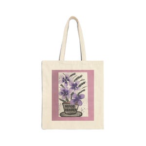May include: A canvas tote bag with a watercolor painting of purple flowers in a pot. The painting is set against a pink background with the words "MOOD READER" on the pot. The bag has a natural, off-white color.