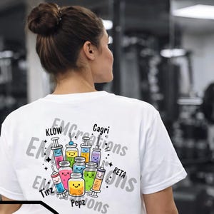 May include: White t-shirt featuring a colorful graphic of cartoon jars and syringes with smiling faces. The design includes the text "EMCreations" and other words. The shirt is worn by a person with their back to the camera.