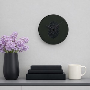May include: A round wall clock with a dark gray face featuring a black pinecone illustration and black hands. The clock is displayed alongside a black vase filled with light purple flowers, a stack of three black books, and a white ceramic mug.