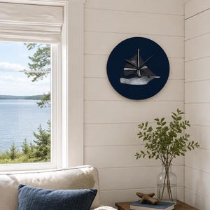 May include: A circular wall clock featuring a dark navy blue background with a stylized illustration of a sailboat in shades of gray and white. The clock has black hands positioned in the center of the boat graphic, mounted on a white wall.