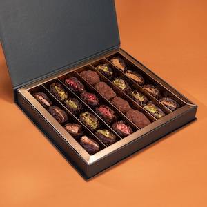 May include: An open, dark gray box with a gold interior holds an assortment of gourmet dates. The dates are filled with various toppings, including pistachios, dried berries, and chocolate. The box sits on an orange background.