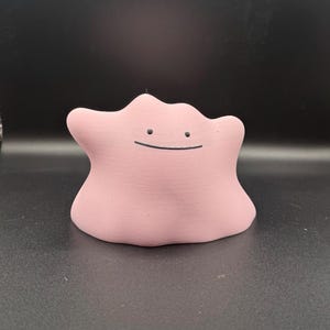 May include: A light pink Ditto figurine with a simple, smiling face. The figure has a rounded, blob-like shape and is set against a dark background. The figurine is a collectible item.