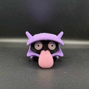 May include: A 3D-printed figure of a cartoon character with a black body, large white eyes, and a pink tongue. The figure has a purple shell-like head with two pointed ears. The figure is on a dark gray surface.