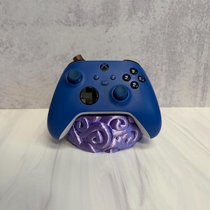 May include: A blue Xbox controller resting on a purple, brain-shaped stand. The controller has black buttons and a black joystick. The stand has a swirling design and is painted in shades of purple.