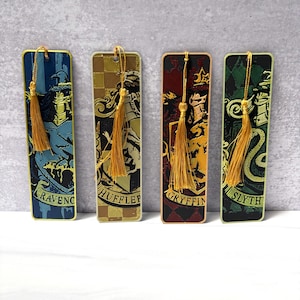 May include: Four rectangular bookmarks with gold tassels. Each bookmark features a different house crest from a fantasy series. The houses are Ravenclaw, Hufflepuff, Gryffindor, and Slytherin. The bookmarks have a gold border and are set against a gray background.