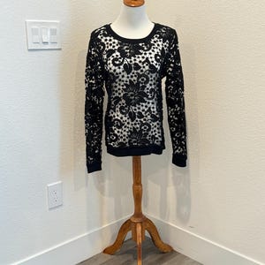 Puede incluir: Top de manga larga de encaje negro con un diseño floral. El top se muestra en un maniquí de madera. El diseño de encaje cubre toda la parte superior, incluidas las mangas. El cuello y los puños son de color negro sólido.