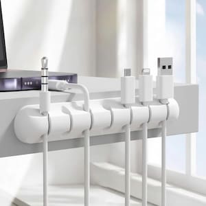 May include: A white cable organizer with multiple slots, holding various white charging cables. The organizer is mounted on a white surface, with a laptop visible in the background. The cables include USB, USB-C, and Lightning connectors.