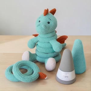 May include: A crocheted teal dragon toy with orange accents, sitting next to two teal rings and two gray cones. The dragon has white feet and a sad expression. One cone has a label that reads "Cone".