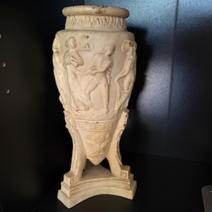 Könnte beinhalten: Eine cremefarbene, dekorative Vase mit detaillierten Flachreliefs von menschlichen Figuren in Bewegung. Das Gefäß hat einen breiten Rand, einen verjüngten Mittelteil und steht auf einem dreibeinigen Sockel mit kunstvollen Verzierungen.