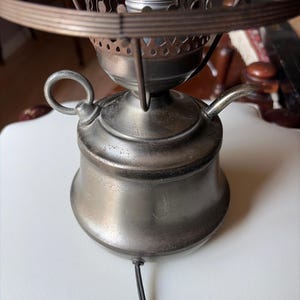 Könnte beinhalten: Ein metallischer, dunkel getönter Lampenfuß mit rundem Körper, einem seitlichen Griff und einem kleinen Ausguss. Die Oberseite verfügt über einen kreisförmigen Metallrahmen und eine zentrale Fassung für eine Glühbirne. Ein schwarzes Kabel führt heraus.