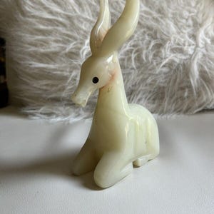 May include: A carved, off-white figurine of a deer in a seated position. The deer has long, curved horns, black eyes, and a smooth, polished surface. The figurine is a decorative item.