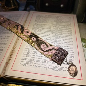 May include: A vintage bookmark with a pink and gold floral design lies on an open book. The bookmark has a decorative metal clasp and a small pendant. The book's pages are aged, with text visible.