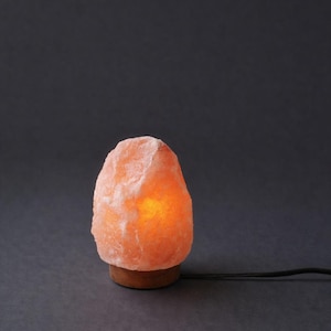 May include: A glowing Himalayan salt lamp. The lamp has a rough, pinkish-orange surface and sits on a small, round wooden base. A black power cord extends from the base. The background is a dark gray.