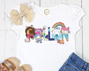 May include: White short-sleeved t-shirt with a colorful graphic of the name "FALLON" spelled out in a playful, patchwork style with cartoon characters. The shirt has a ruffled hem and is paired with denim shorts and sandals.