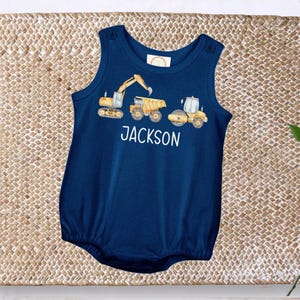 May include: Navy blue baby romper with a construction vehicle design. The romper features a yellow excavator, dump truck, and road roller. The name "JACKSON" is printed in white below the vehicles. The romper has snap closures on the shoulders.