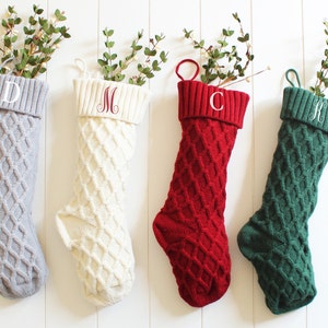 May include: Four cable-knit Christmas stockings in gray, cream, red, and green, each with a different embroidered initial. The stockings are filled with sprigs of greenery and hang against a white wooden background.