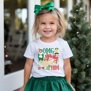May include: A young child wearing a white t-shirt with the text "OMG Santa I Know Him" in red, green, and yellow. The child is also wearing a green skirt and a green bow in their hair. A Christmas tree is in the background.