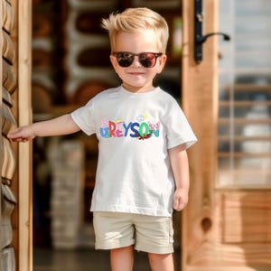 May include: A young child wearing sunglasses and a white t-shirt with the name "Greyson" in colorful letters. The shirt also features cartoon graphics. The child is wearing khaki shorts.