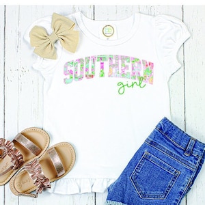 May include: White t-shirt with a pink and green floral design that says "Southern girl" on it. The shirt has a ruffled hem and is paired with denim shorts.