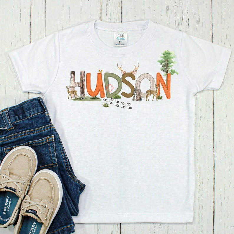 May include: White t-shirt with a woodland-themed design featuring the name "HUDSON" in orange, green, and brown letters. The letters are decorated with deer, trees, and other woodland elements.
