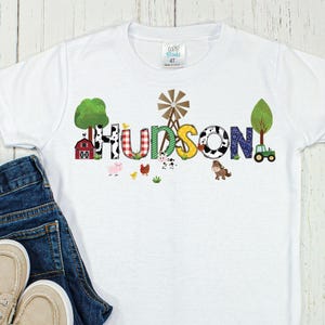 May include: White t-shirt with a farm-themed design featuring the name "HUDSON" in colorful letters with farm animals and a windmill.