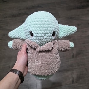 May include: A handmade crochet plush toy of a character with large ears and a brown coat. The toy is primarily light green with black eyes and a brown coat. The plush is soft and textured.