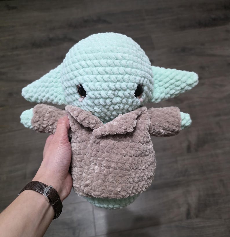 May include: A handmade crochet plush toy of a character with large ears and a brown coat. The toy is primarily light green with black eyes and a brown coat. The plush is soft and textured.