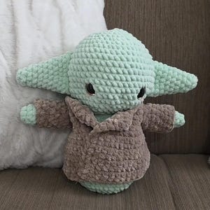 May include: A crocheted plush toy in the likeness of a character with large ears. The toy is primarily light green with a brown robe. The toy has black eyes and is sitting on a brown surface.